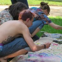 Link to Art page. [Image description: Three people are crouched down in the grass on a sunny day and painting on a long strip of paper.]